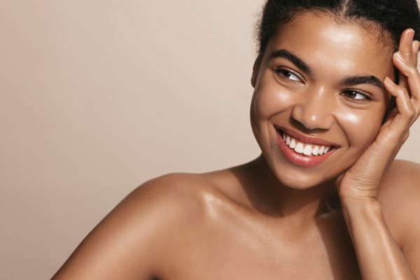 Smiling young woman with glowing facial skin, touching her perfect nourished face, brown background.