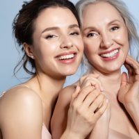 Happy smiling cheerful excited satisfied grey-haired senior mother and brunette young daughter holding hand and hugging with love tenderness closeup studio portrait. Different age generation bonding