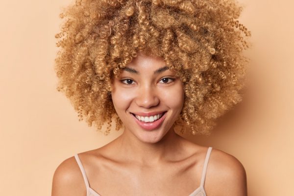 Close up portrait of positive curly haired woman smiles gently has silky healthy skin no make up poses bare shoulders looks directly at camera isolated over beige background. Beauty concept.